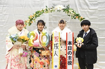 写真：振袖姿の女性3人とスーツ姿の男性1人が、20の形の風船や祝成人、本日の主役と書かれたタスキを持って記念撮影をする様子