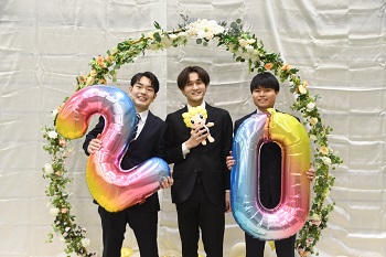 写真：スーツを着た男性3人が、20の形の風船やざまりんのぬいぐるみを持って記念撮影をする様子