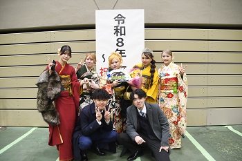 写真：令和8年座間市成人式～祝生誕20年～と書かれた立て看板の前で記念撮影をする振袖姿の女性5人とスーツ姿の男性2人