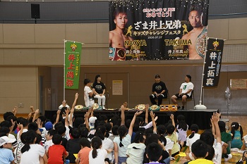 写真：いのうえ兄弟への質問コーナーの様子。壇上には、左から市長、いのうえなおやさん、いのうえたくまさんがおり、挙手をする子どもたちが映っています。
