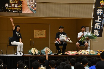 写真：勝利報告の様子。壇上には左から市長、いのうえなおや選手・たくま選手が座っており、お二人が獲得したベルトが飾られています。