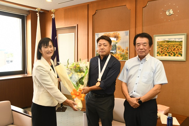 写真:(左から)花束を掲げる市長とまんとく てつのりさん、くらや まもるさん が並ぶ集合写真