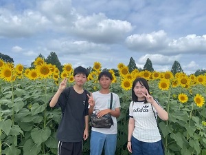 写真：国際親善大使がひまわ畑でピースをして写っている
