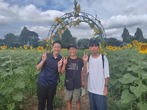 写真：国際親善大使がひまわり畑でピースをして写っている