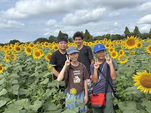 写真：国際親善大使4名がひまわり畑の中こちらを向いて写っている